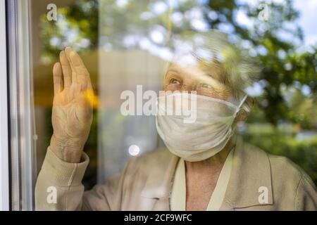 Ältere Frau trägt eine schützende Gesichtsmaske und blickt aus dem Fenster Zu Hause Stockfoto