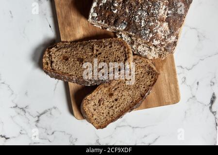 Draufsicht auf leckeren gesunden Vollkorn Roggenbrot Laib mit Scheiben auf Holzschneidebrett auf Marmortisch serviert Stockfoto