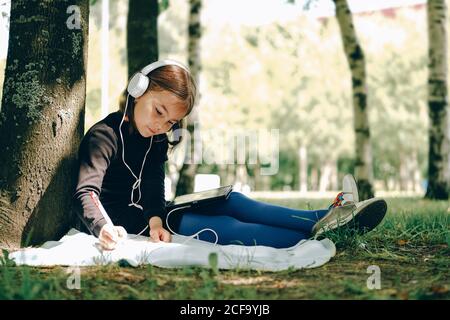 Glückliches Mädchen in weißen Kopfhörern mit digitalen Tablet-pc im Park. Fernlernkonzept. Resilienz, zurück zur Schule, neue Normalität. Stockfoto