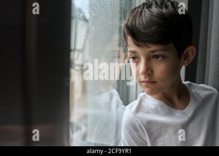 Gelangweiltes Kind in lässiger Kleidung im Wohnzimmer und stehen Lehnt sich an das Fenster und beobachtet den Regen auf der Straße Stockfoto