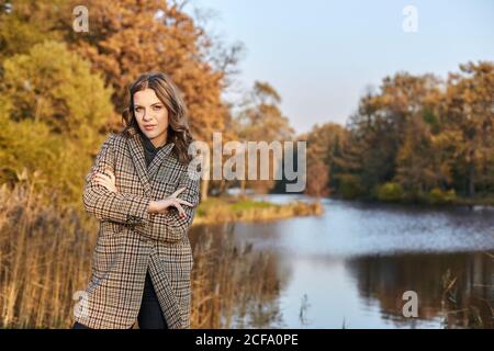 Ernste langhaarige Frau trägt braunen Mantel und steht im Herbst park an einem See Stockfoto