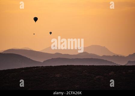 Silhouetten entfernter Heißluftballons, die gegen den Sonnenuntergang fliegen Über hügeliges Gelände Stockfoto