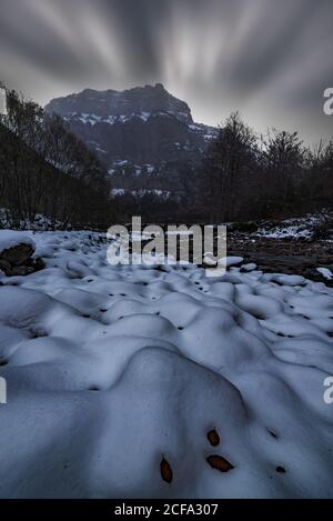 Schmaler kleiner Bach in der Mitte von dünnen Bäumen, die laufen Durch das teilweise mit Schnee bedeckte Bergtal unter wolkenbedecktem Himmel Bei Einbruch der Dunkelheit im Winter Stockfoto