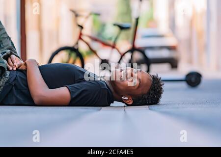 Seitenansicht des glücklichen Jugendlichen afroamerikanischen Mannes, der auf liegt Asphaltstraße in der Straße Blick auf die Kamera Stockfoto