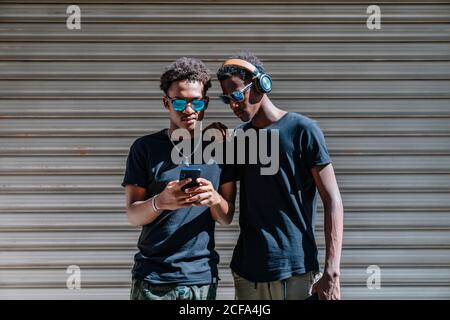 Cool jugendlich afroamerikanischen männlichen Teenagern in Sonnenbrillen fotografieren Mit Mobiltelefon, während Sie im Sonnenlicht auf der Straße stehen Stockfoto