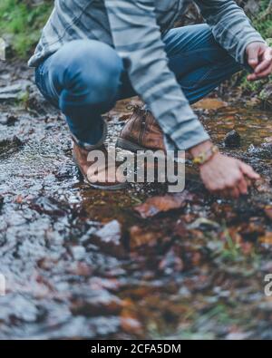 Von unten der Ernte männlich trägt grauen Pullover und Jeans Mit braunen Stiefeln, die während des Spazierens auf nassem Pfad hocken Herbstwald Stockfoto