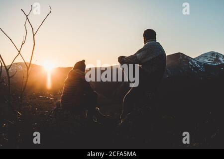 Rückansicht eines entspannten Mannes und einer Frau, die hinten auf der Bergspitze sitzen und den wunderschönen Sonnenuntergang mit verschneiten Hügeln im Hintergrund genießen Stockfoto
