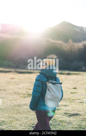 Rückansicht des unerkennbaren jungen Reisenden in warmer aktiver Kleidung Mit Rucksack und Sonnenbrille überqueren leere Trockenrasenfeld in Herbstsonniger Tag mit Wald und Bergen im Hintergrund Stockfoto