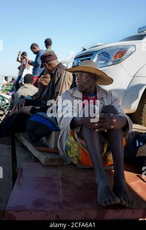 Madagaskar - 6. JULI 2019: Barfuß schwarzer Mann in Hut und Aboriginal-Outfit sitzt auf verwitterten Brettern von geparkten Auto neben lässigen Menschen bei Tageslicht Stockfoto