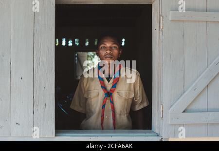 Madagaskar - 6. JULI 2019: Seriöser schwarzer Mann in Uniform mit buntem Schal, der am Fenster mit hölzernen Fensterläden steht Stockfoto