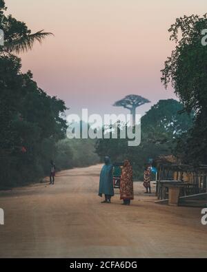 Madagaskar - 6. JULI 2019: Menschen in bunten Umhänge stehen an staubigen Straße durch ländliche tropische Dorf und Wald bei Sonnenuntergang Stockfoto