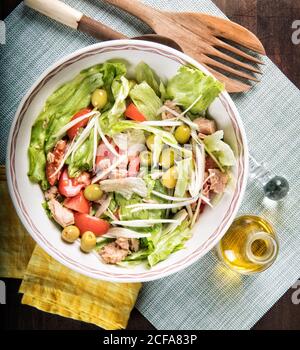 Von oben gesund lecker Salat mit frischen Tomaten und Salat mit grünen Oliven und Huhn in einer Schüssel serviert Tisch mit Holzlöffel und Flasche Olivenöl Stockfoto