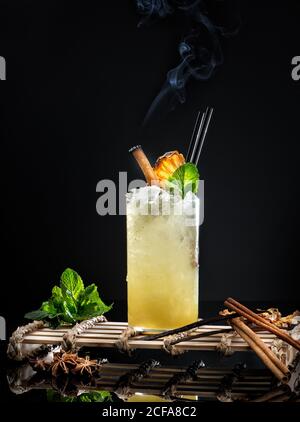 Ein Glas gelbes, würziges alkoholisches Getränk, garniert mit frischer Minze Blatt und Rauchen Zimt Stick vor schwarzem Hintergrund Stockfoto