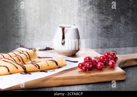 Leckere hausgemachte Pfannkuchen oder Crepes mit Schokoladensirup serviert Holzbrett mit frischen roten Beeren gegen schäbige Wand Stockfoto