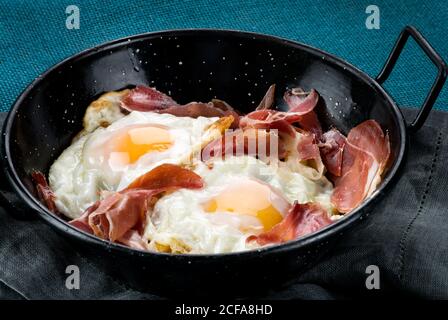 Pfanne mit Spiegeleiern mit traditionellem spanischen iberischen Schinken serviert Auf rustikalem Holztisch mit Küchenutensilien und Produkten Stockfoto