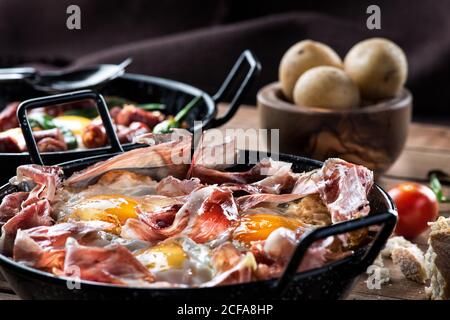 Pfanne mit Spiegeleiern mit traditionellem spanischen iberischen Schinken serviert Auf rustikalem Holztisch mit Küchenutensilien und Produkten Stockfoto