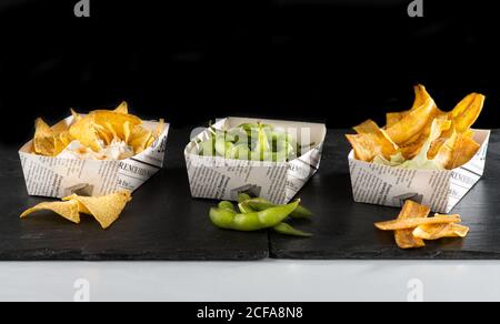 Ein großer Winkel von leckeren mexikanischen Snacks wie knackige Nachos Und edamame mit gebratenen Bananen in Papierschüsseln auf dunkel Holztisch vor schwarzem Hintergrund im Restaurant Stockfoto