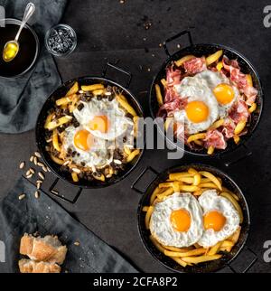 Draufsicht auf Bratkartoffel und Eier in schwarz rund Pfannen mit geröstetem Schinken oder Blutwurst in der Zusammensetzung Mit Brot und Zutaten auf dem Tisch in der Küche Stockfoto