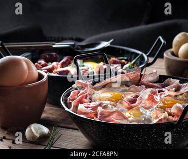 Pfanne mit Spiegeleiern mit traditionellem spanischen iberischen Schinken serviert Auf rustikalem Holztisch mit Küchenutensilien und Produkten Stockfoto