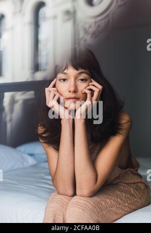 Blick durch das Fenster einer jungen charmanten Brünette Frau, die in die Kamera schaut und im Schlafzimmer auf dem Bett sitzt Stockfoto
