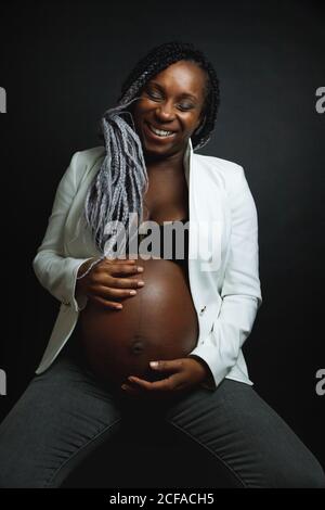Junge fröhlich schwangere afroamerikanische Frau mit Zöpfen tragen weiß blazer und Jeans streicheln den Bauch, während sie im Studio dagegen sitzen Schwarze Wand Stockfoto