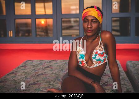 Nachdenklich ruhig afroamerikanische Frau trägt bunte Bikini-Top und Kopfband sitzt in der Lounge im Zimmer mit rotem Boden und Roter Sonnenuntergang durch Fenster im Hintergrund Stockfoto