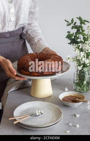 Anonyme Frau in Schürze Putting-Teller mit frischem Bundt Kuchen Auf dem Tisch neben Blumen und Apfelsoße Stockfoto