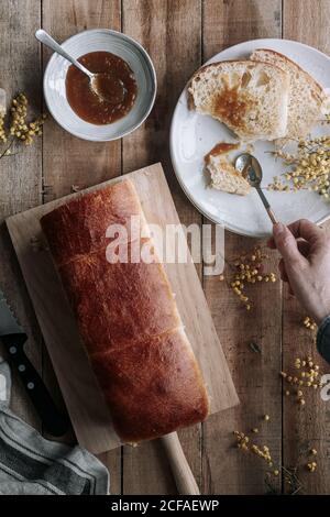 Von oben beschnittene unerkennbare Personenhand mit Rechteck Laib frisches Brioche Brot auf Holztisch mit Schüssel Von Marmelade und Messer Stockfoto