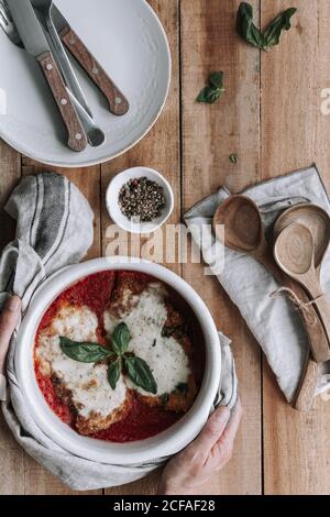 Draufsicht auf anonyme Person setzen heiße Schüssel voll von Leckeres Huhn mit Parmesan und Basilikumblättern in Tomate geröstet Sauce auf Holztisch neben Gewürzen und Geschirr Stockfoto