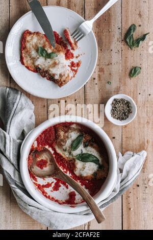 Draufsicht auf Schüssel und Teller mit leckeren Huhn mit Käse und Basilikum in Tomatensauce in der Nähe Serviette gelegt geröstet Und Gewürze auf Holztisch Stockfoto