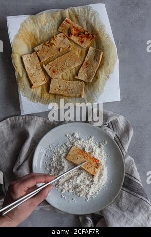 Draufsicht der Ernte anonym mit Zero Waste Essstäbchen und Tofu-Scheiben in Sojasauce auf den Teller geben Mehl steht auf weißem Küchentuch auf grauem Tisch Stockfoto