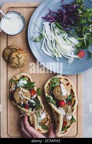 Oben anonyme Ernte Person Hände auf Lebensmittelzusammensetzung falafel Und frisches Gemüse auf Brot in der Nähe mit Salat auf Schneiden Platine Stockfoto