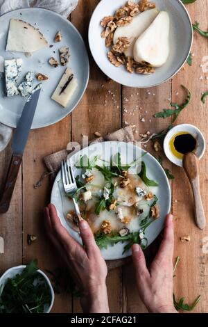 Draufsicht auf anonyme Person Putting Teller mit Birnensalat Mit Rucola auf Holztisch in der Nähe von Käse und Walnüssen Küche Stockfoto