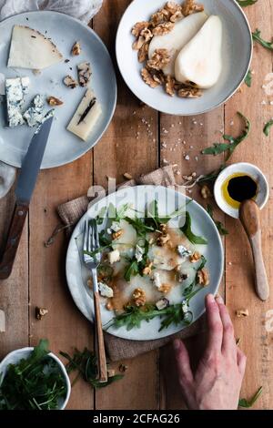 Draufsicht auf anonyme Person Putting Teller mit Birnensalat Mit Rucola auf Holztisch in der Nähe von Käse und Walnüssen Küche Stockfoto