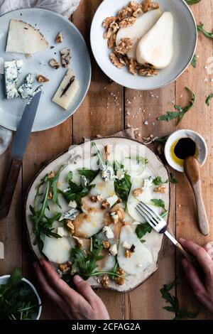 Draufsicht auf anonyme Person Putting Teller mit Birnensalat Mit Rucola auf Holztisch in der Nähe von Käse und Walnüssen Küche Stockfoto