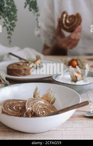 Von oben von geschnittenen leckeren hausgemachten süßen Brötchen Kuchen mit Schlagsahne und Physalis getrocknete Blumen auf dem Teller serviert Holztisch mit Zutaten Stockfoto