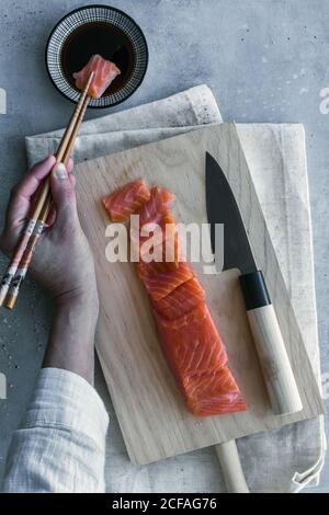 Von oben Ernte anonyme Person hält Stück Lachs mit Essstäbchen und Dipping in Sauce am Tisch serviert Stockfoto