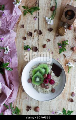 Draufsicht auf Scheiben von frischen Kiwi und Himbeeren platziert In der Nähe Schokolade und Haselnüsse in Schüssel mit gesunden Brei auf Holztisch in der Nähe Fliedertuch Stockfoto