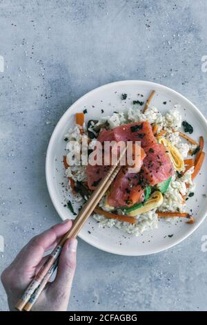 Von oben beschnittene Hand einer nicht erkennbaren Person, die Holzstäbchen hält Zu essen lecker appetitlich geschnittener Lachs auf weißem Reis mit Gemüse auf dem Tisch Stockfoto