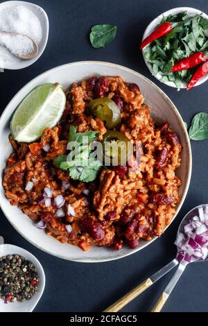 Draufsicht auf traditionelle scharfe Chili mit Fleisch und Bohnen Garniert mit Limette und Petersilie mit mariniertem grünem Pfeffer serviert In weißer Schüssel auf Tisch mit Zutaten Stockfoto