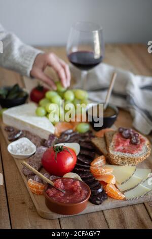 Von oben gesichtslose zugeschnittene Person Hände essen Snacks aus Holz Tablett mit Fleischgemüse Obst und Glas Rotwein Stockfoto