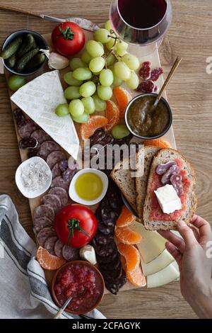 Von oben gesichtslose zugeschnittene Person Hände essen Snacks aus Holz Tablett mit Fleischgemüse Obst und Glas Rotwein Stockfoto