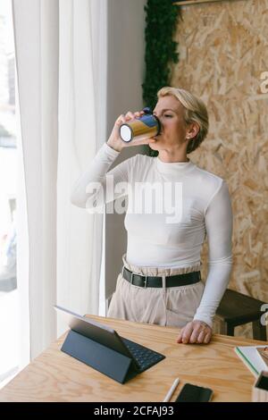 Verträumte stilvolle blonde Frau am Holztisch mit Tablet und Papeterie, die einen Schluck Kaffee aus der Einweg-Tasse nimmt und im Büro wegschaut Stockfoto