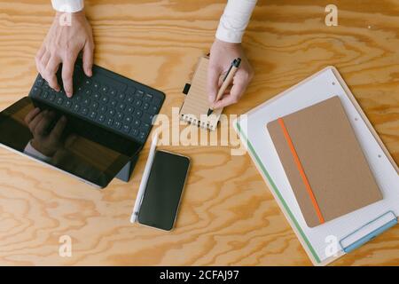 Schneiden Sie die Draufsicht der Frau, die auf dem Tablet-Tastenfeld tippt, während Sie Notizen in einem kleinen Notizblock auf einem Holztisch machen Stockfoto