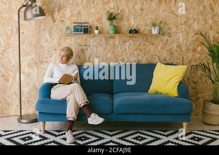 Blonde Frau mit kurzen Haaren in weißem Hemd sitzt auf dem Sofa und schaut weg und schreibt in einem Notizbuch, das an einem Geschäftsprojekt arbeitet Stockfoto