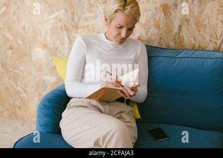 Blonde Frau mit kurzen Haaren in weißem Hemd, die auf dem Sofa sitzt und in einem Notizbuch schreibt, das an einem Geschäftsprojekt arbeitet Stockfoto