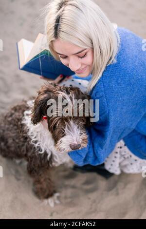 Von oben glücklich junge Frau in lässigen Pullover und Rock Halten Buch und streicheln liebenswert lockigen Hund, während Ausgaben kostenlos Zeit am Sandstrand Stockfoto