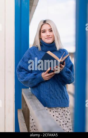 Nachdenklich junge Frau in warmen Pullover und Rock lächelnd und Blick auf die Kamera, während auf Holzzaun mit offenen lehnt Buch gegen weiß-blau gestreifte Wand Stockfoto