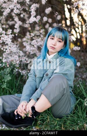 Traurige Millennial weibliche Modell mit blauen Haaren in stilvollem Outfit Betrachten Sie die Kamera nachdenklich, während Sie auf grünem Gras in der Nähe sitzen Blühender Baum im Frühlingsgarten Stockfoto