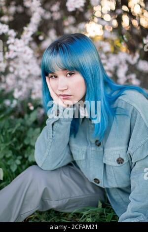 Traurige Millennial weibliche Modell mit blauen Haaren in stilvollem Outfit Betrachten Sie die Kamera nachdenklich, während Sie auf grünem Gras in der Nähe sitzen Blühender Baum im Frühlingsgarten Stockfoto